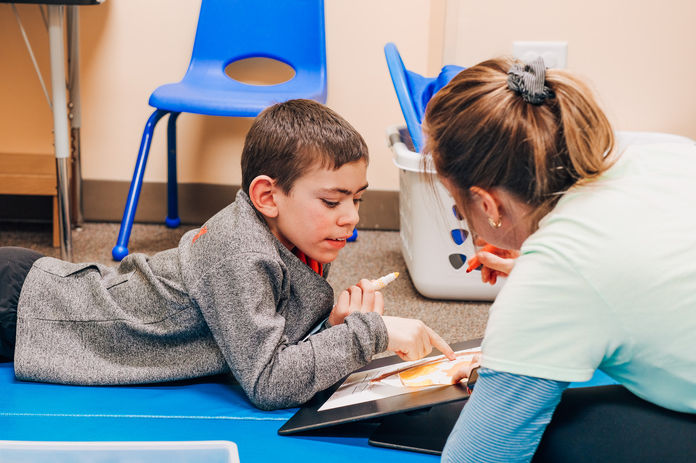An occupational therapist on the floor during a therapy session with a young boy. He is pointing to a piece of paper with one hand and has a marker in the other hand. They are both looking down at the paper.