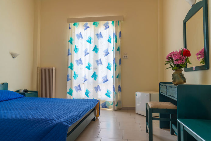 Bedroom with blue bed, curtain, and dressing table stay in Aegina Hotel.
