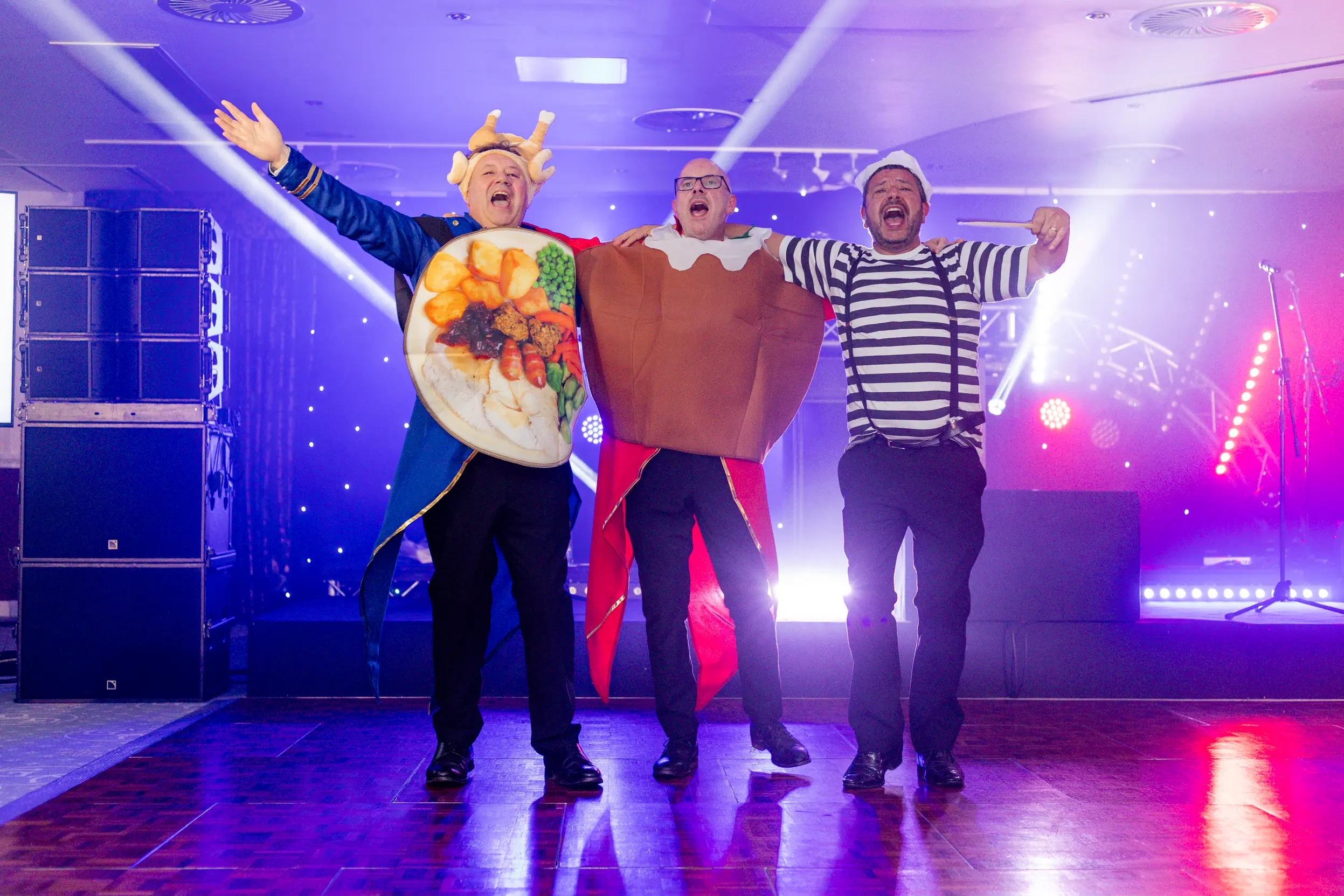 Danny and Tom Lillington with Gary Hewitt dressed in humorous costumes, including a roast dinner, a Christmas pudding, and a mime, performing joyfully on stage during the Paris-themed party.