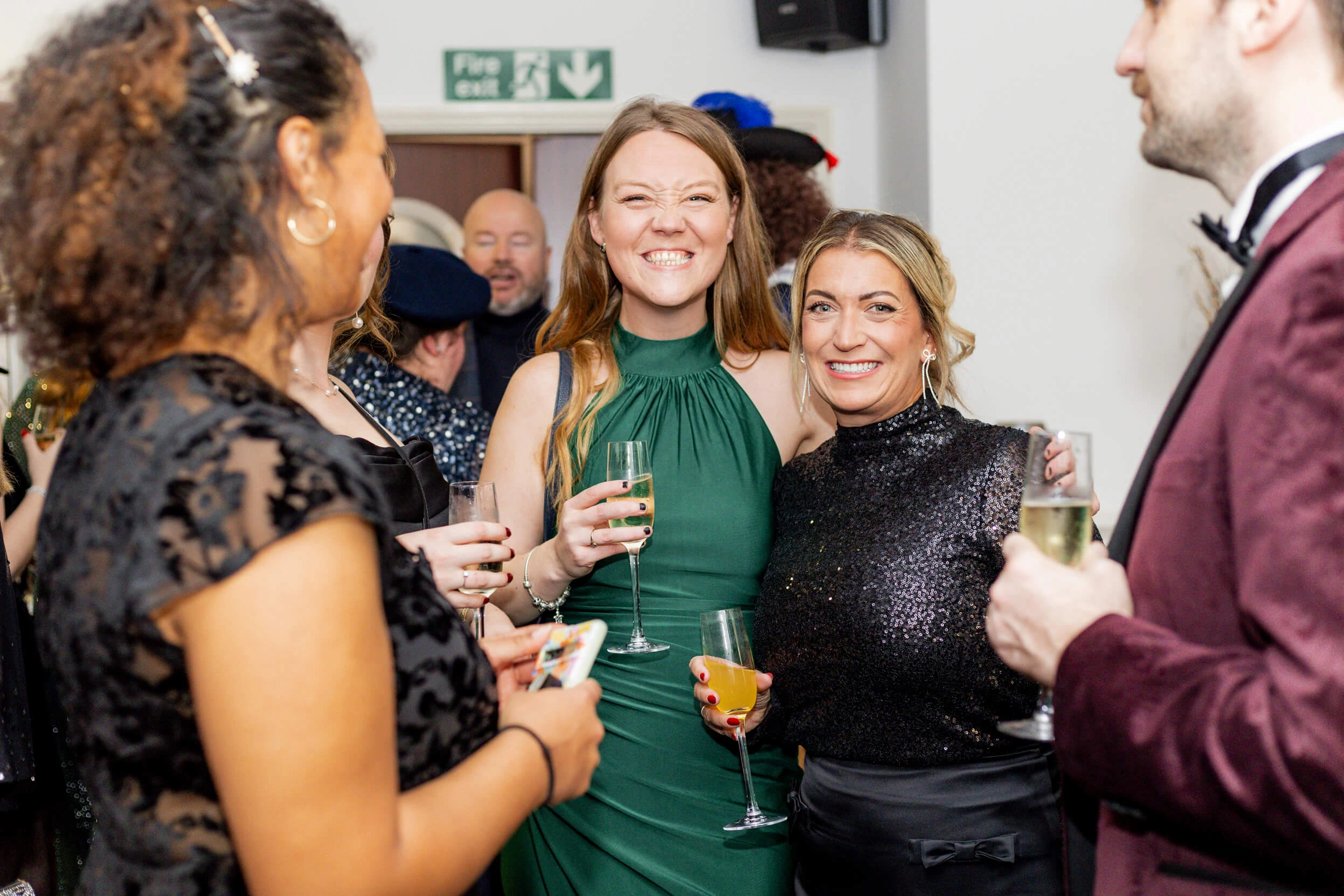 Two women posing for a photo, dressed in elegant evening attire, with other attendees mingling in the background.