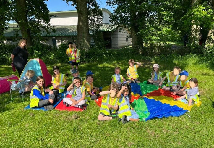 Children wearing high-visibility jackets sitting on picnic blankets outside on a sunny summer day, enjoying an outdoor gathering.