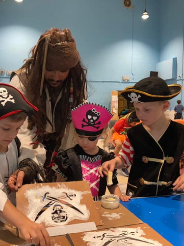 A pirate helping children decorate pizza boxes with skeleton designs, guiding them through the creative process while they have fun adding spooky details for Halloween.