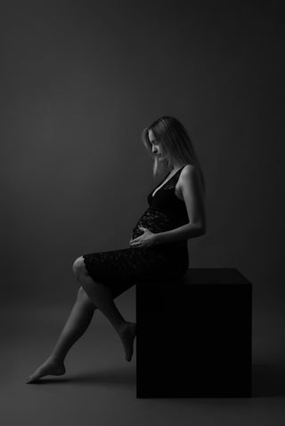 אישה הריונית יושבת על קובייה שחורה בסטודיו , עם שמלה תחרה, , מבט לבטן ההריונית שלה.