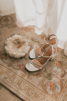 Sandalias de tacón blancas co tiras de pedrería junto a un tocado floral.