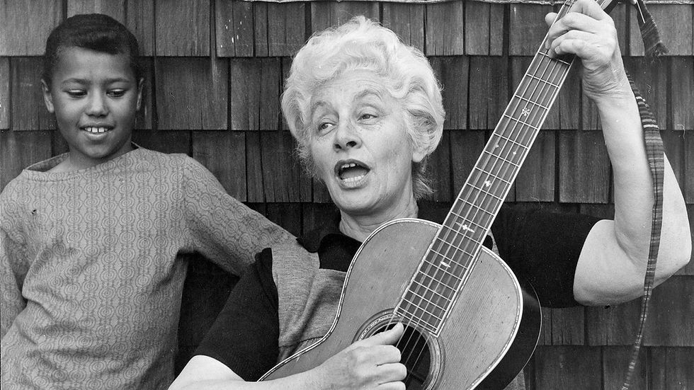 A white-haired Caucasian singer-songwriter, Malvina Reynolds, cradles her guitar over her left shoulder while playing in 1970. An African American boy stands next to Malvina focusing on her guitar playing fingers.