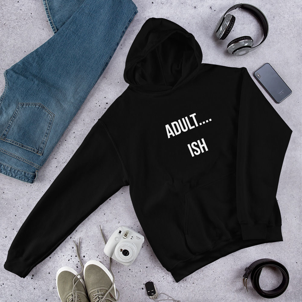 Black unisex hoodie laid flat with white text that reads “ADULT…ISH”, styled with jeans, trainers, and headphones.
