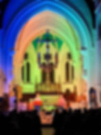Interior of St Nicolas' Church in Guildford looking towards the altar and chancel. Two musicians (Ninebarrow) are performing in front of the altar. They are standing and playing keyboard instruments and singing into microphones. The walls and ceiling of the church are lit with multicoloured light