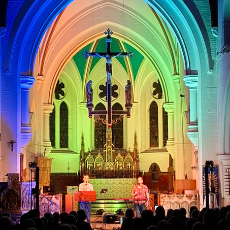 Interior of St Nicolas' Parish Church in Guildford. View towards the altar and chancel. Two musicians (Ninebarrow) are standing in front of the altar, performing on a harmonium and electronic keyboard and singing into microphones. The walls and ceiling are lit with multicoloured lights ranging from purples and blues to green, orange and yellow. A large audience is sitting watching them.