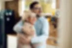 A joyful older woman with short blonde hair and glasses is warmly embraced by a younger man in a light blue shirt. Both are smiling broadly, standing in a sunlit kitchen with warm wood and white tones. The moment conveys comfort, reassurance, and the restored relationships that can follow a fair resolution supported by a retrospective mental capacity assessment.