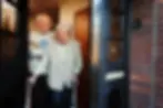 An older couple stands warmly in the doorway of their home, smiling at the camera. The man, wearing glasses and a patterned jumper, stands slightly behind the woman, who also wears glasses and leans on a walker for support. The open door and welcoming expressions convey approachability and trust, while subtly reflecting situations — such as wills, property sales, or healthcare choices — where a retrospective mental capacity assessment might be required to review decisions made in the past.