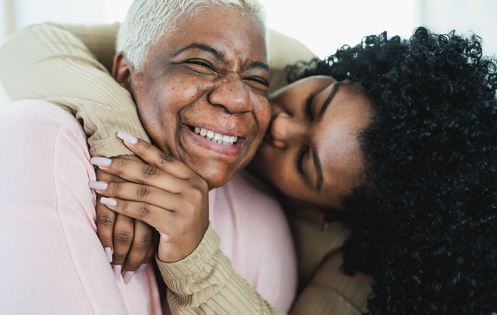Certainly. In the image, there are two individuals sharing a close and affectionate moment. On the left, there's an older woman with short, white hair, laughing heartily. She is wearing a beige sweater and her face radiates joy. On the right, there's a younger woman with curly, dark hair. She is closely embracing the older woman, her face pressed against the older woman's cheek, seemingly giving her a gentle kiss. The younger woman's hand, adorned with white nail polish, is placed gently on the older woman's face. The atmosphere of the image conveys a strong bond, warmth, and happiness between the two individuals.