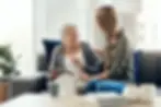 An older woman with short grey hair sits comfortably on a modern grey sofa, smiling warmly at a younger woman with a long braid. They are seated close together, leaning in as they look at a tablet device in the older woman’s hands. The scene is bright and welcoming, with natural light from a nearby window, soft blue cushions, and indoor plants creating a relaxed atmosphere. The interaction conveys empathy, guidance, and trust — reflecting the supportive nature of retrospective mental capacity assessments.