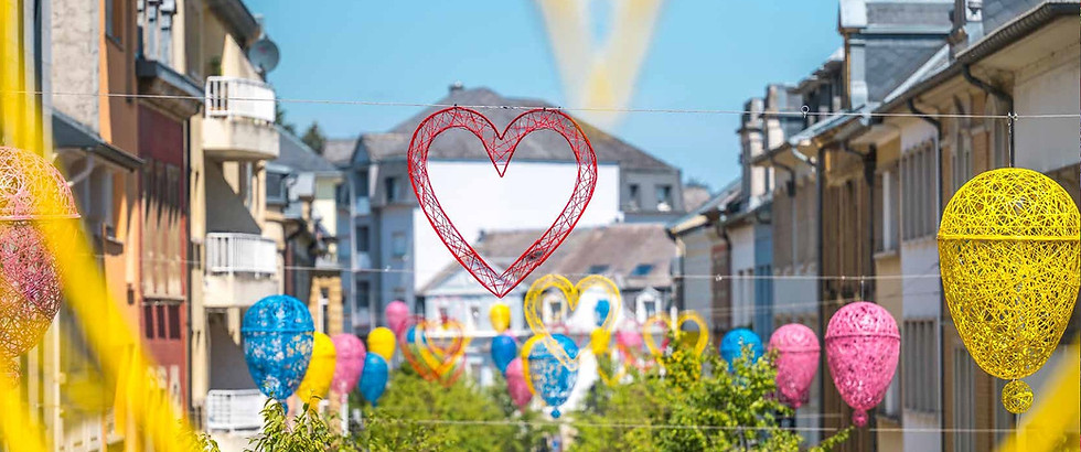 Auf dem Bild sieht man eine festlich geschmückte Innenstadt mit farbenfroher Saison-Dekoration. Über der Strasse hängen dekorative Elemente aus geflochtenem Material in Form von Herzen und Luftballons. Die Farben – Gelb, Blau, Pink und Rot – erzeugen eine lebendige, fröhliche Atmosphäre, ideal für den Sommer oder besondere Anlässe wie Stadtfeste, Märkte oder Themenwochen. Die Outdoor-Dekorationen sind an Drahtseilen über der Strasse befestigt und ziehen sich durch die gesamte Einkaufsstrasse.  Diese Art von Strassen-Dekoration eignet sich perfekt für temporäre saisonale Installationen in Innenstädten, Einkaufszonen oder auf öffentlichen Plätzen. Die Kombination aus leichten Materialien, wetterfestem Design und kreativer Farbgestaltung macht die Dekoration nicht nur optisch ansprechend, sondern auch funktional und langlebig – ideal für den Einsatz im Visual Merchandising im Aussenbereich.