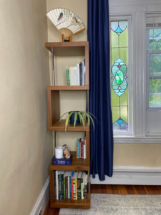 A modern wooden bookshelf with a unique zigzag design stands in the corner of a room. It contains various books, a plant, and decorative items including a folded fan. A navy blue curtain and stained glass window are visible in the background.