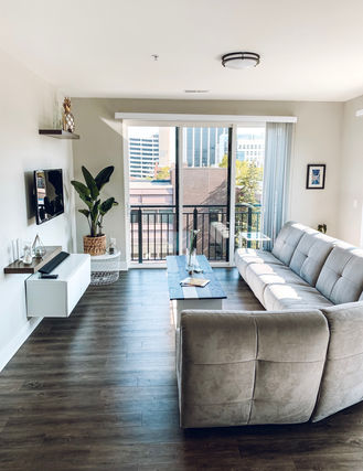 A modern living room with a light gray sofa facing a wall-mounted TV. A wooden coffee table sits in front of the sofa. Large glass doors open to a balcony with a city view. The room has light walls, hardwood floors, and various plants for decor.