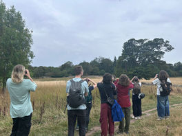 Six people are facing away from the camera in a field edged with trees. They are all looking through binoculars. It is a cloudy day.