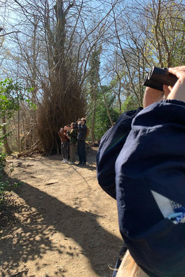 In the foreground is someone's arms holding binoculars up to a face we cannot see. Behind them are four people looking in the same direction through their binoculars. They are all in the woods on a dry dirt path.