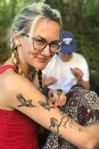 El is has brown and blonde hair in two plaits. They are wearing tortoiseshell glasses, a red tank top and red patterned trousers. She is holding a blue tit in their hand and showing the camera the bird next to their blue tit tattoo on their right arm.