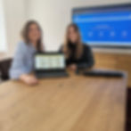 Zwei Mitarbeiterinnen sitzen an einem großen Holztisch im Besprechungsraum und schauen grinsend und gespannt in die Kamera. Vor ihnen liegt ein Laptop, auf dem der Ticketshop des Hunsrücker Oktoberfestes angezeigt wird. Im Hintergrund ist der Bildschirm des Besprechungsraumes zu sehen, auf dem der Countdown zum Start des Vorverkaufs läuft, der in knapp 2 Stunden endet.