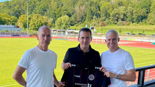 Drei Männer stehen auf der Tribüne eines Fußballplatzes und halten ein schwarzes Trikot in der Hand.