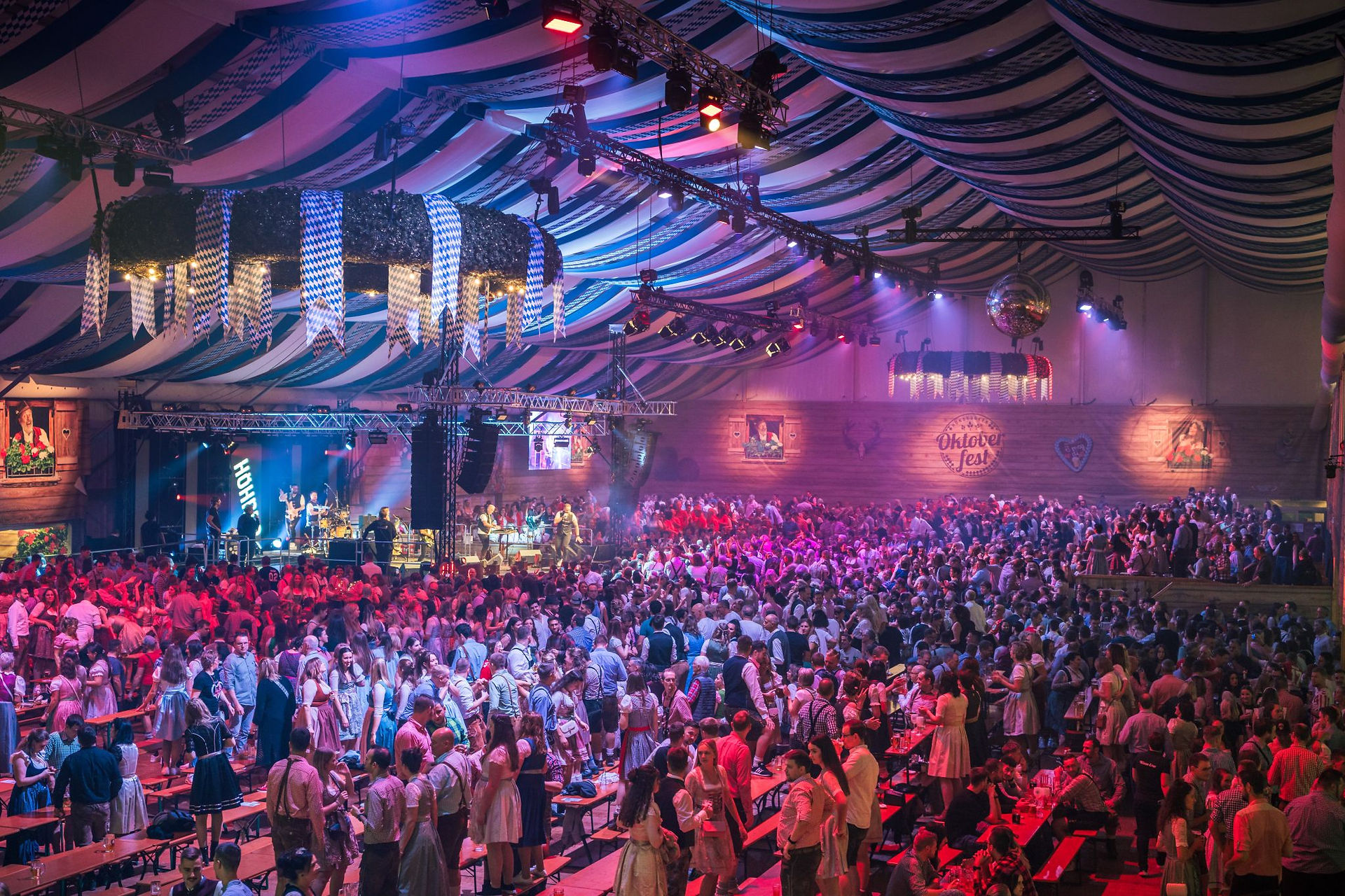 Ein voll besetztes Festzelt beim Hunsrücker Oktoberfest: Unzählige Menschen in Tracht stehen feiernd auf Bierzeltgarnituren. Im Hintergrund ist die Bühne zu sehen, auf der eine Band live auftritt.