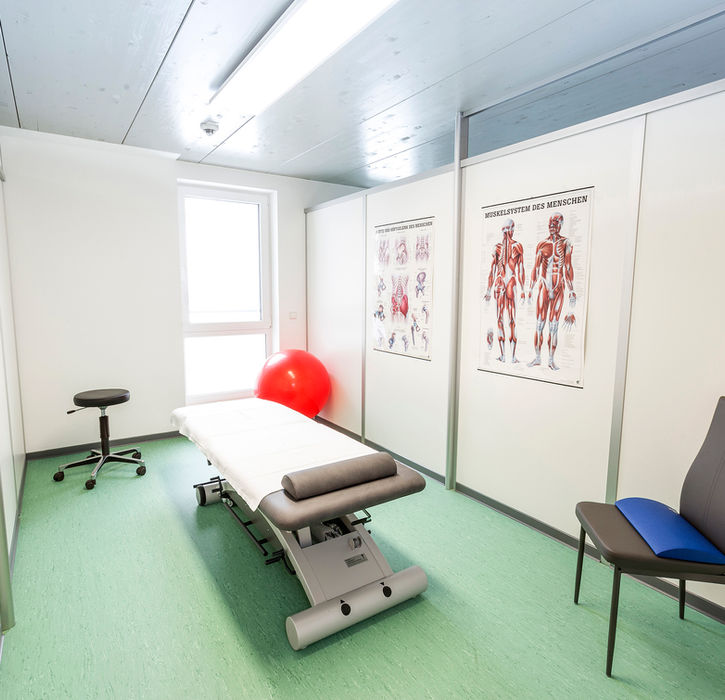 Medizinischer Behandlungsraum mit Anatomieplakat, Untersuchungsliege und Stuhl, hell eingerichtet.