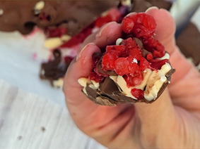 Hand holds chocolate-covered raspberries with white and dark chocolate drizzles, set on a light surface. Bright red berries are prominent.