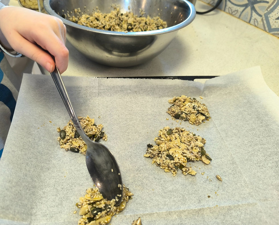A hand spreads seed mix with a spoon onto parchment paper. A metal bowl with more mix is visible in the background. Bright and clear setting.