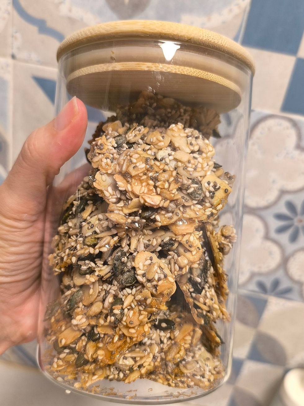 Hand holds a glass jar filled with mixed seeds and nuts against a patterned blue and beige tiled background. Mood is natural and rustic.