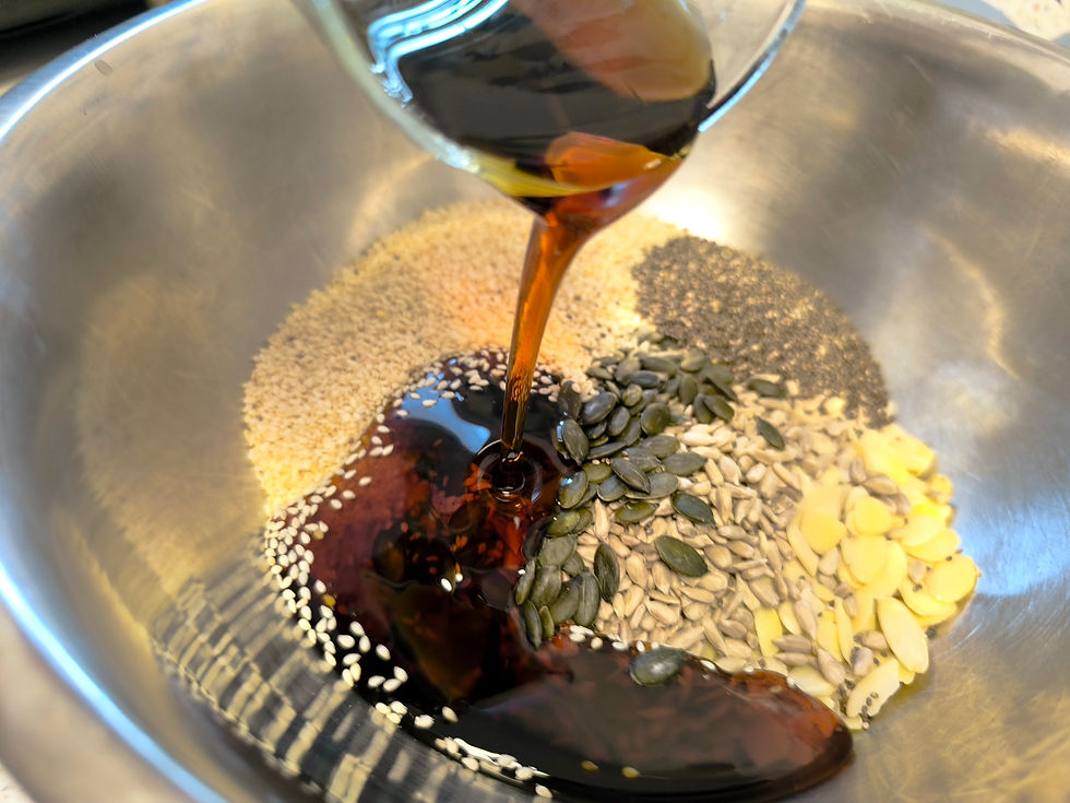 Seeds and nuts in a metal bowl with dark syrup being poured over. Mix includes sesame, sunflower, pumpkin seeds. Warm kitchen setting.