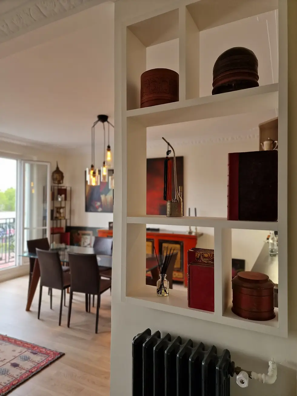 Vue traversante depuis l'entrée au travers d'une bibliothèque sur mesure blanche ajourée servant de séparateur d'espace. Ses niches exposent des objets d'art (boîtes laquées, livres) et laissent entrevoir le salon lumineux avec une table à manger en verre, des chaises contemporaines et un grand tableau abstrait rouge et noir au mur.