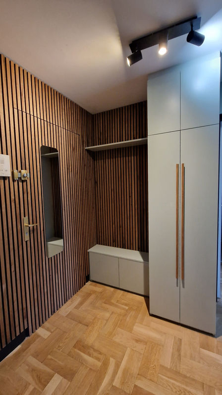 Vue d'ensemble d'une entrée d'appartement rénovée. Dressing sur mesure gris avec poignées en bois, mur décoratif en claustra de bois et éclairage par spots noirs sur rail. Décoration intérieure contemporaine et chaleureuse.