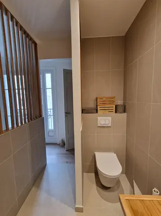 Optimisation d'une petite salle de bain avec WC suspendu et rangements intégrés. Utilisation d'un claustra en bois pour séparer l'espace sans occulter la lumière provenant de la chambre.