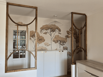 Détail d'une verrière en bois sur mesure au design organique avec motifs en demi-cercles, séparant l'entrée du séjour. En arrière-plan, papier peint panoramique PaperMint à motif d'arbres et buffet blanc épuré, réalisation Lise Dupin-Denisart.