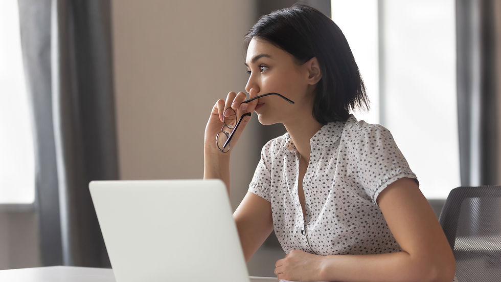 Woman in a dotted shirt, seated at a desk with a laptop, gazes thoughtfully out a window, holding glasses to her lips. Bright, neutral room.