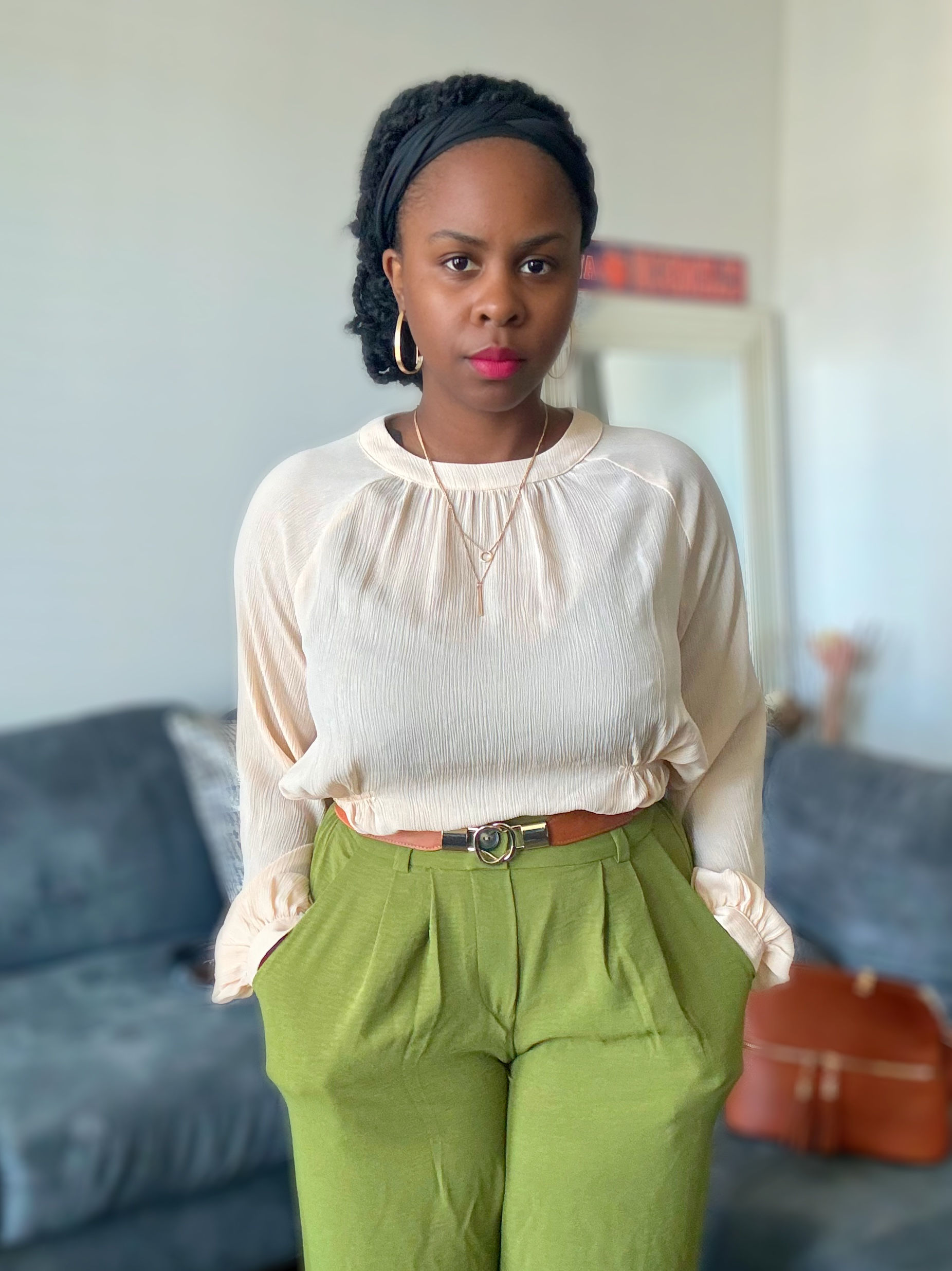 Woman wearing beige top and green pants, posing indoors with a couch.