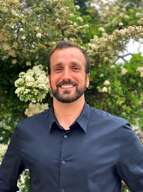 A portrait of Andrea Pescosolido dressed in a black bottomed up shirt, green background with plants and flowers.