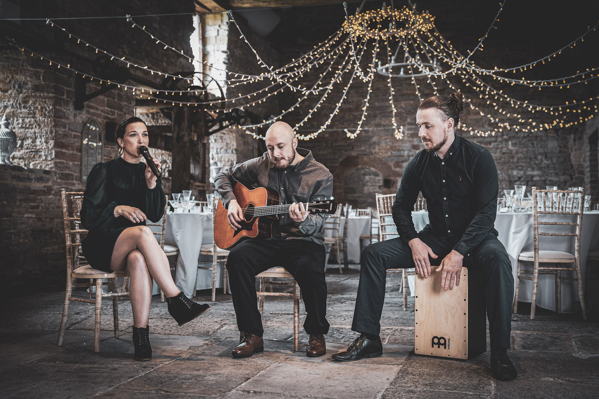 An acoustic trio performing in a rustic wedding venue, featuring a singer, guitarist, and percussionist beneath fairy lights