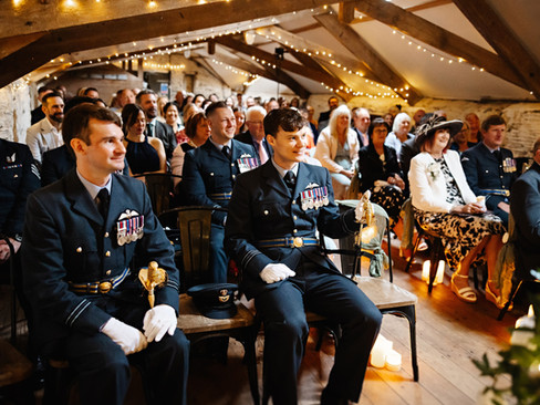 Wedding guests seated inside a warmly lit barn. Groomsmen in RAF uniforms waiting at the front beneath wooden beams and fairy lights