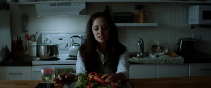 Film still from If I Told You: a woman prepares lobster at a kitchen counter at night, a glass of white wine beside her.