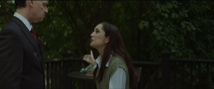 Film still from If I Told You: a woman pleads with a man in a dark suit on a leafy garden terrace at dusk.