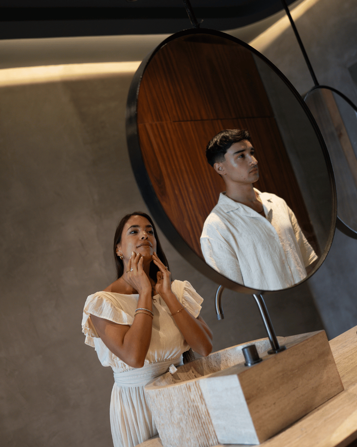 A couple at a sculptural stone vanity in a Maiim suite in Tulum, the man reflected in a round wood-framed mirror.