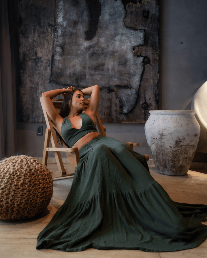 A woman in a flowing green dress reclines in a wooden lounge chair beside a clay urn at Maiim, Tulum.