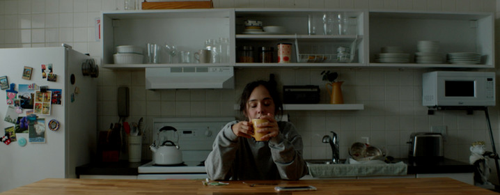 Film still from If I Told You: a woman sits at a kitchen table with her head in her hands, a yellow mug in front of her.