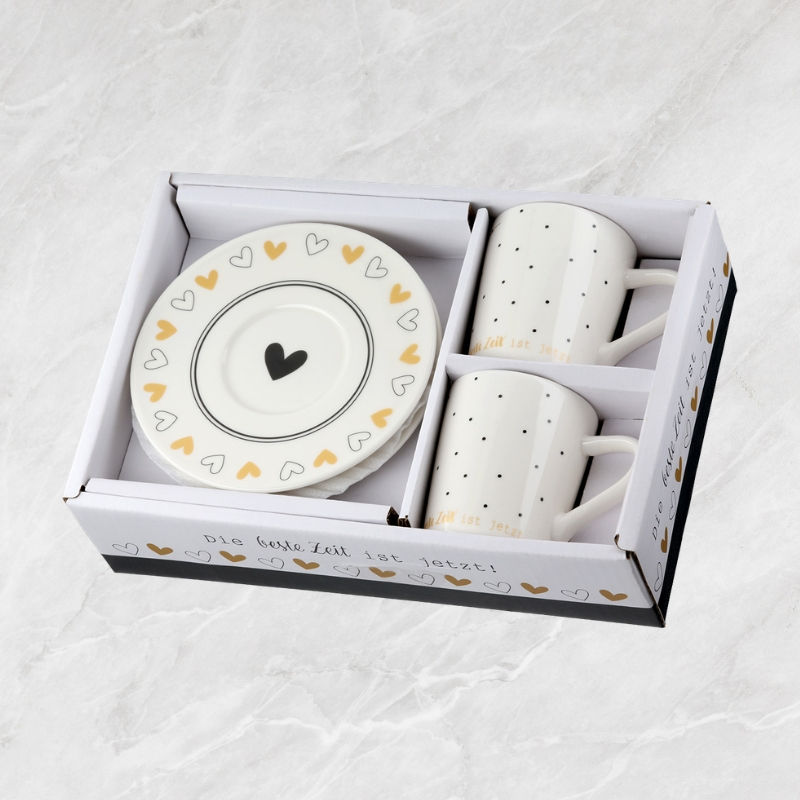 Zwei weiße Espressotassen mit schwarzen Punkten, goldenem Schriftzug und Untertassen mit Herzmotiven in Karton.