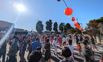 Large crowd enjoying outdoor festival under hanging lanterns on a sunny day.