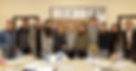 A group of 11 people smiling and standing close together indoors. Behind them are Japanese characters on a wall. Water bottles and plates are on the table. The atmosphere appears friendly and warm.