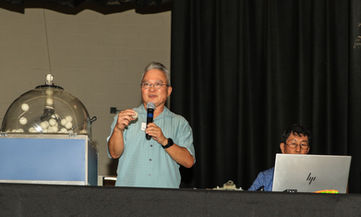 Man announcing raffle numbers from machine while another man uses laptop.