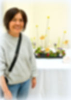 A person in a gray sweater smiles beside an artistic flower arrangement on a table. The colorful flo
