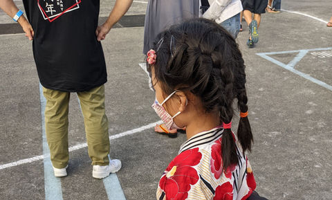 Young girl in red floral yukata, pigtails, mask, outdoor setting.